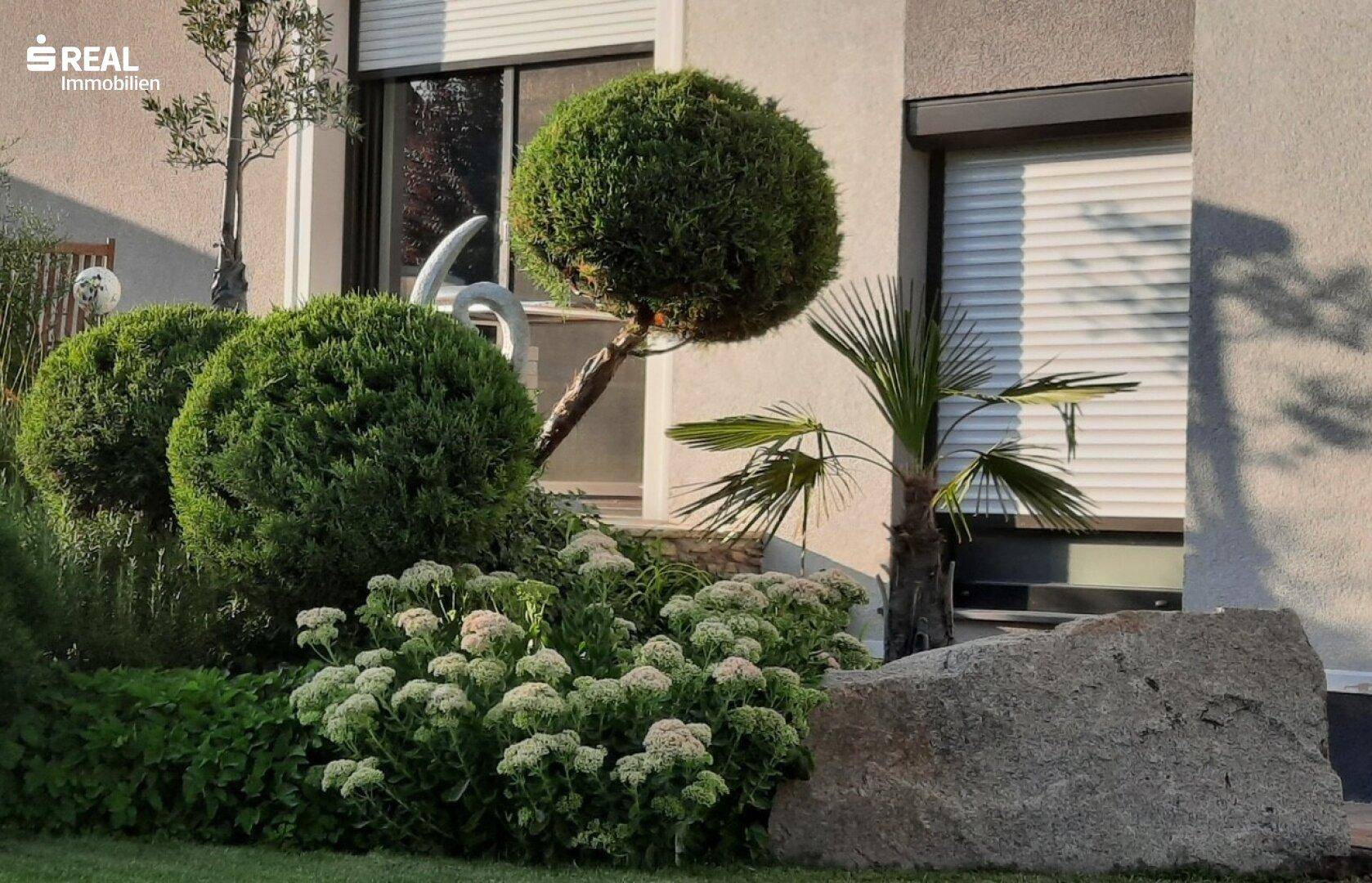 Gepflegter Gartenbereich mit Ziersträuchern, Blumen und einem großen Stein vor der Hausfassade.