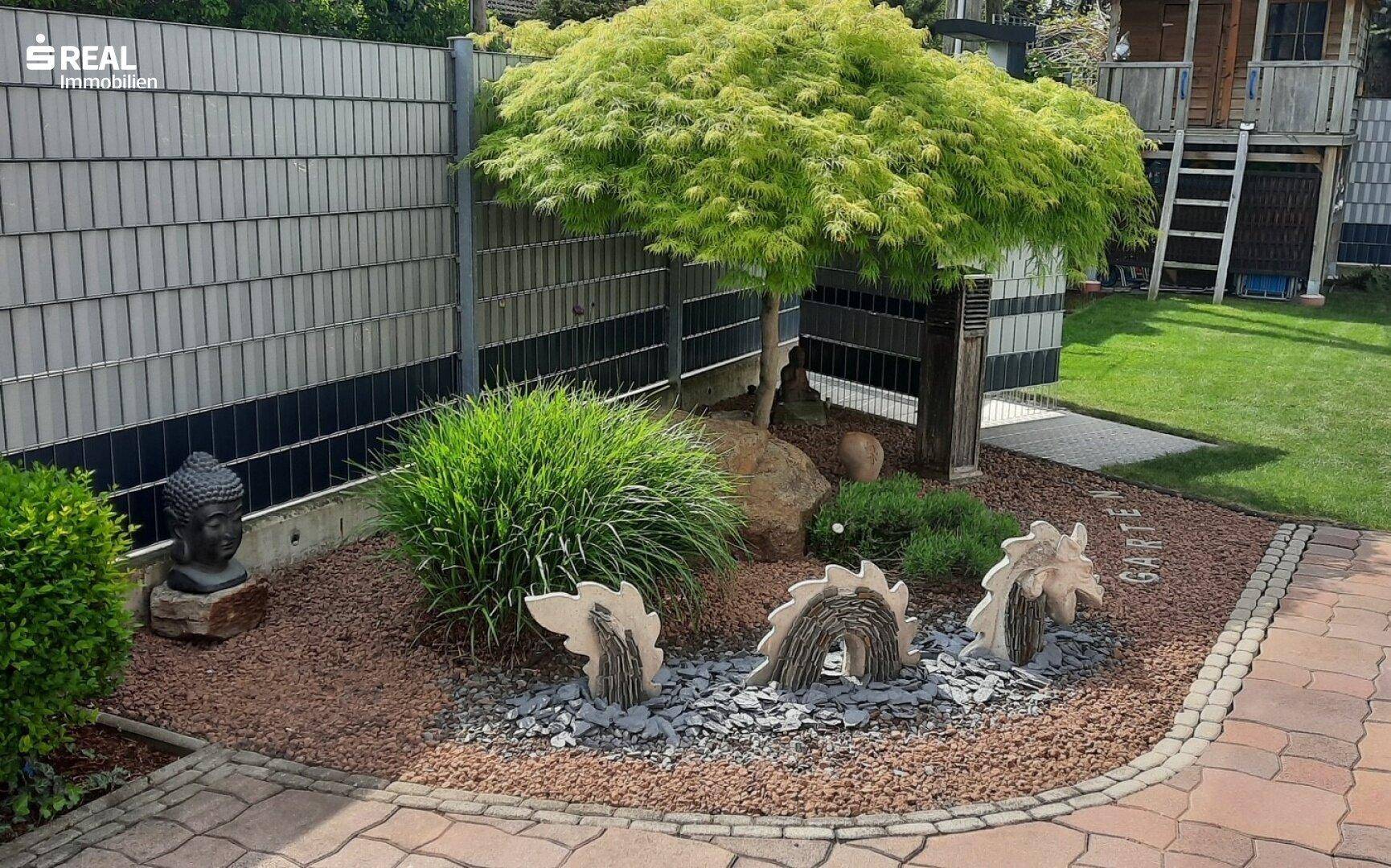 Mediterran gestalteter Gartenbereich mit Ahornbaum, dekorativen Steinen und Buddha-Statue.