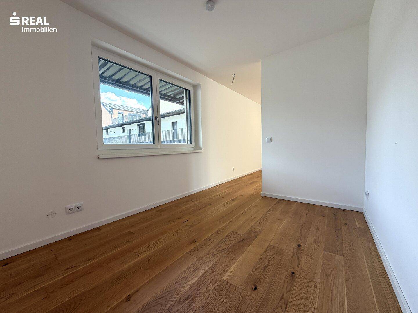 Leeres Zimmer mit Holzboden, weißen Wänden und einem Fenster mit Blick nach draußen.