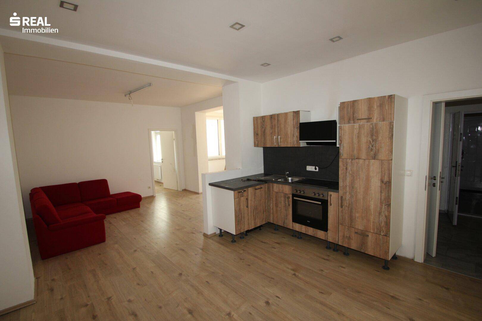 Offener Wohnbereich mit Holzboden, rotem Sofa und einer Küchenzeile mit Holzfronten und integriertem Herd.