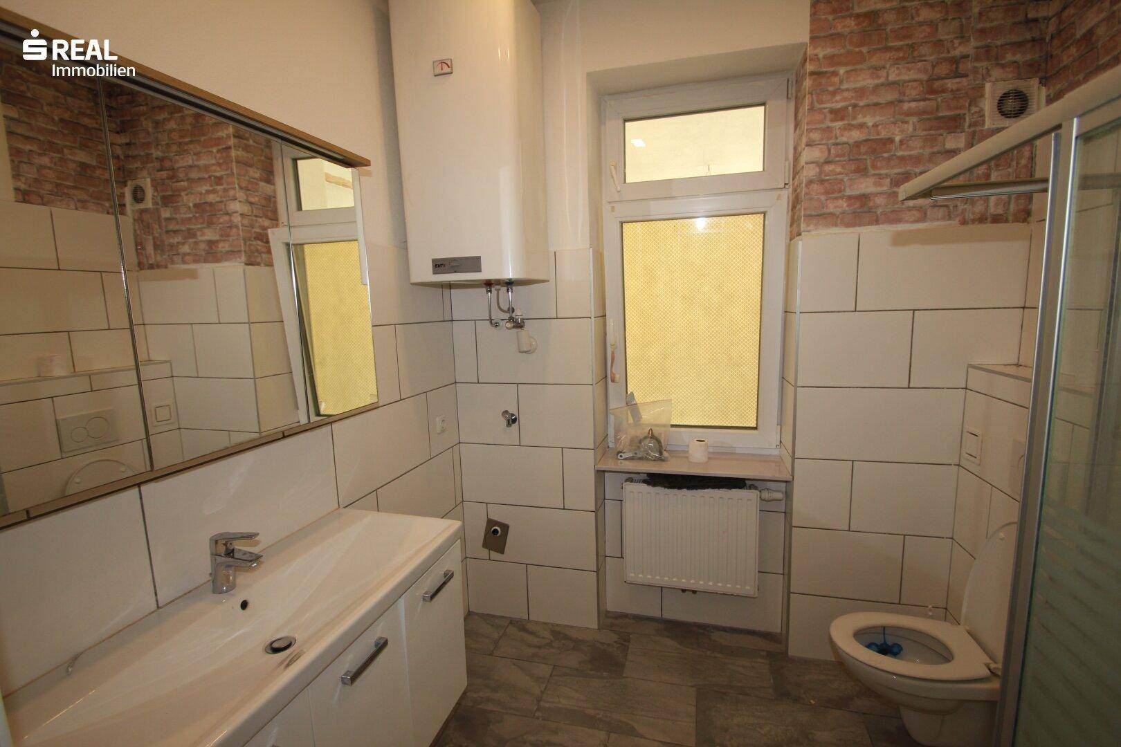 Badezimmer mit weißen Fliesen, Duschkabine, WC, Waschtisch mit Unterschrank und einem Fenster.