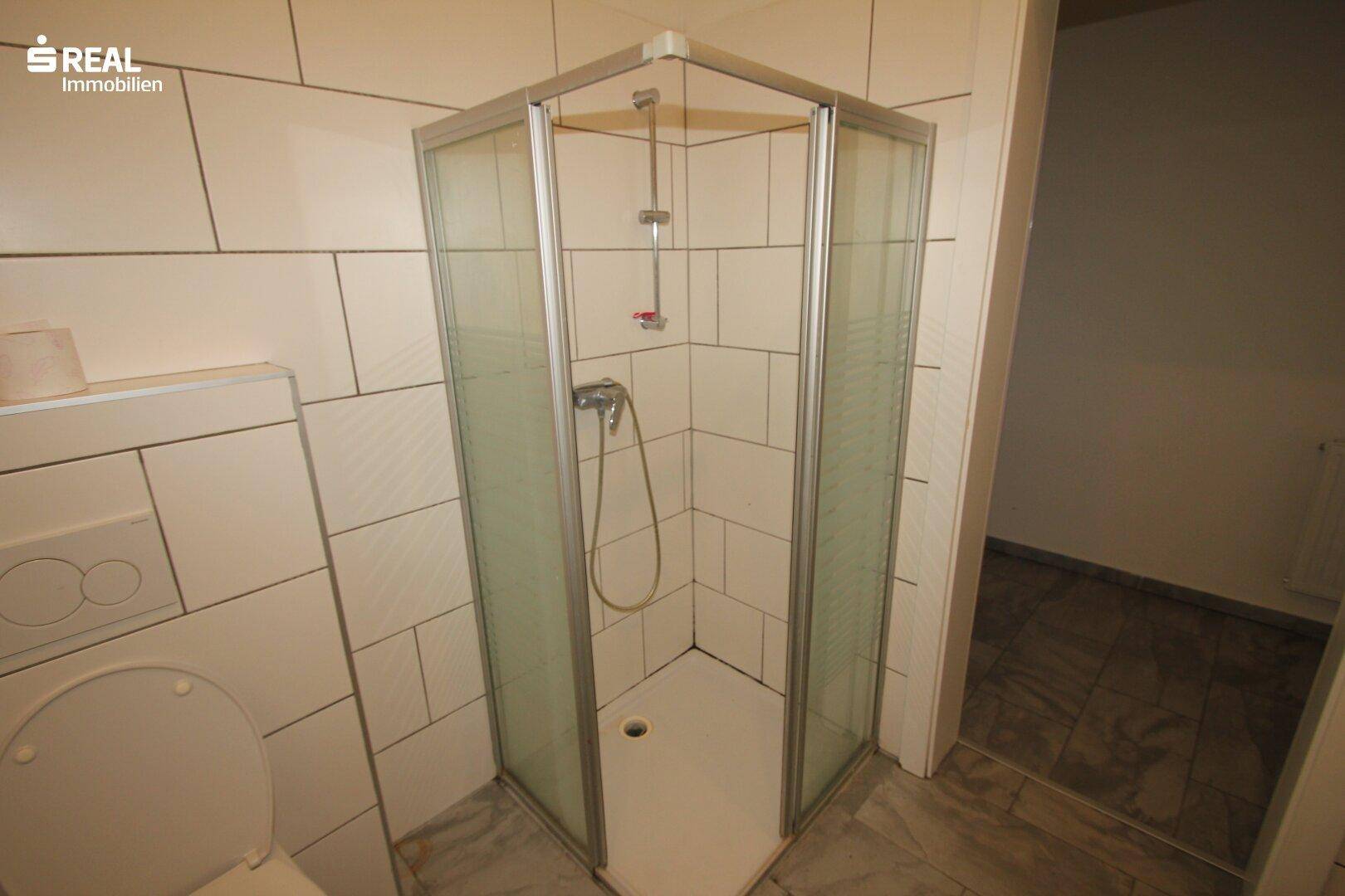 Duschkabine in einem gefliesten Badezimmer mit WC und dunklem Boden, bereit zur Nutzung.