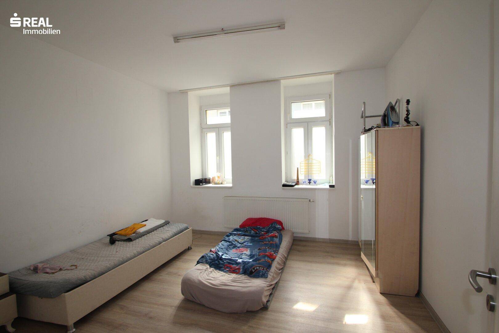 Schlafzimmer mit Holzboden, zwei Einzelbetten und einem Kleiderschrank, hell durch zwei Fenster.