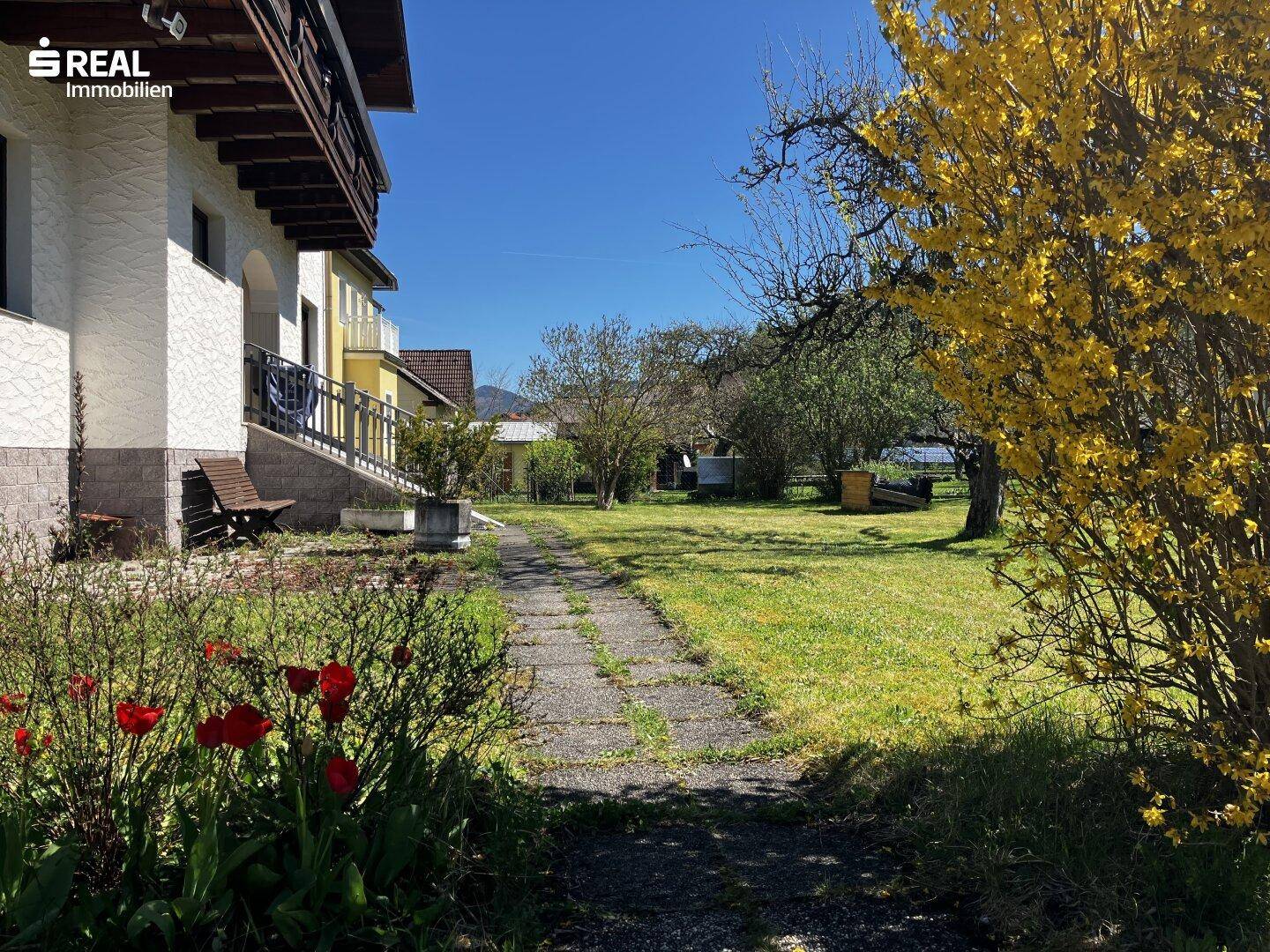 Großer Gartenbereich mit Rasenfläche, Blumenbeeten und einem Weg, umgeben von Bäumen.
