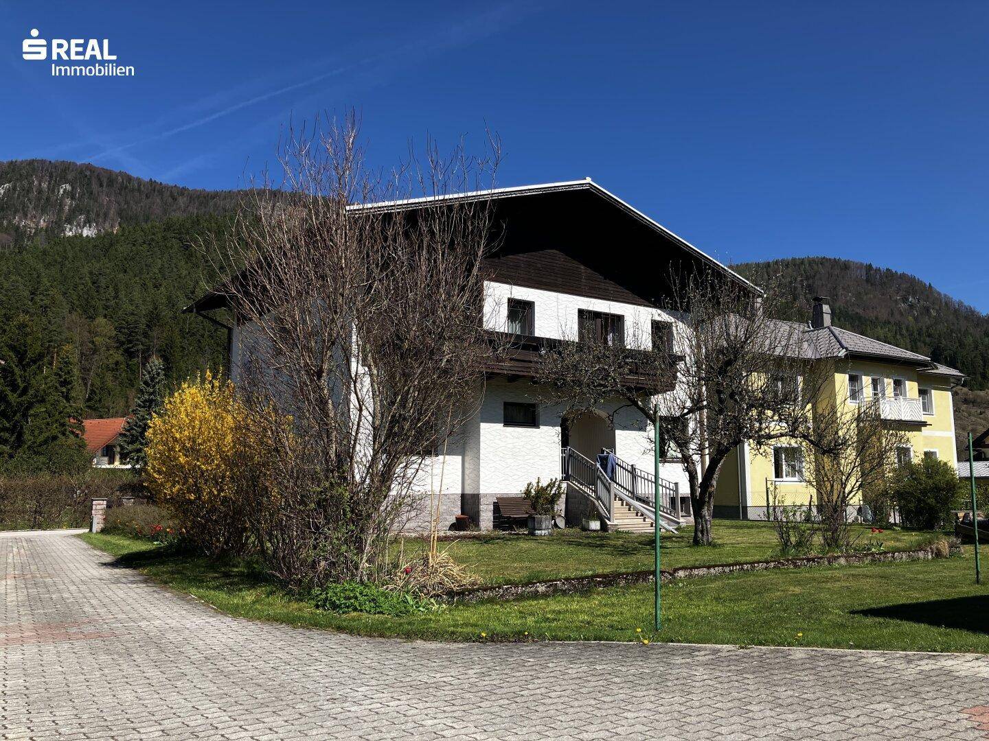 Seitenansicht des Hauses mit Zufahrt und Blick auf die umliegende Berglandschaft an einem sonnigen Tag.