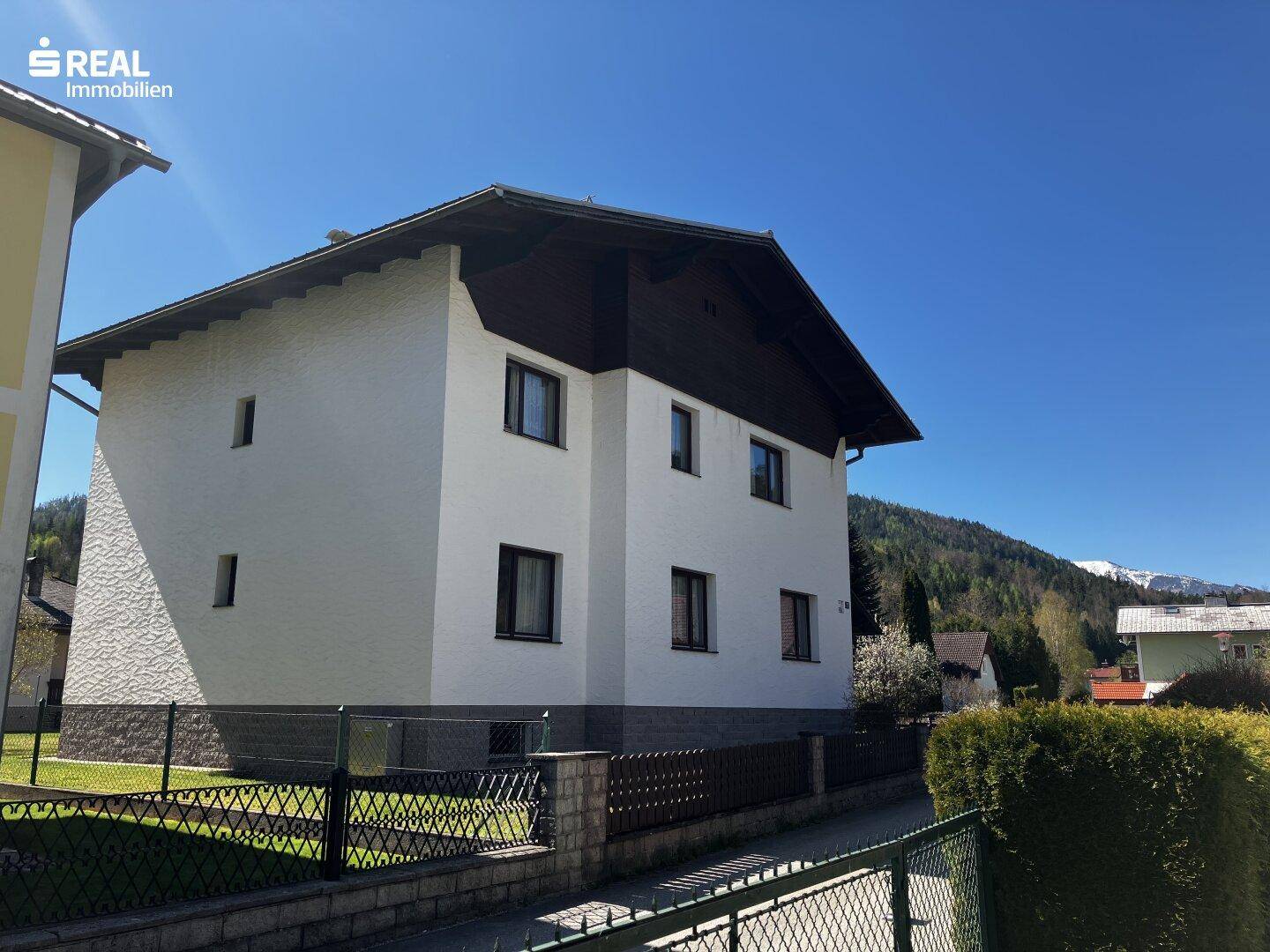 Seitenansicht des weißen Einfamilienhauses mit Zaun und Blick auf die grüne Umgebung und Berge.