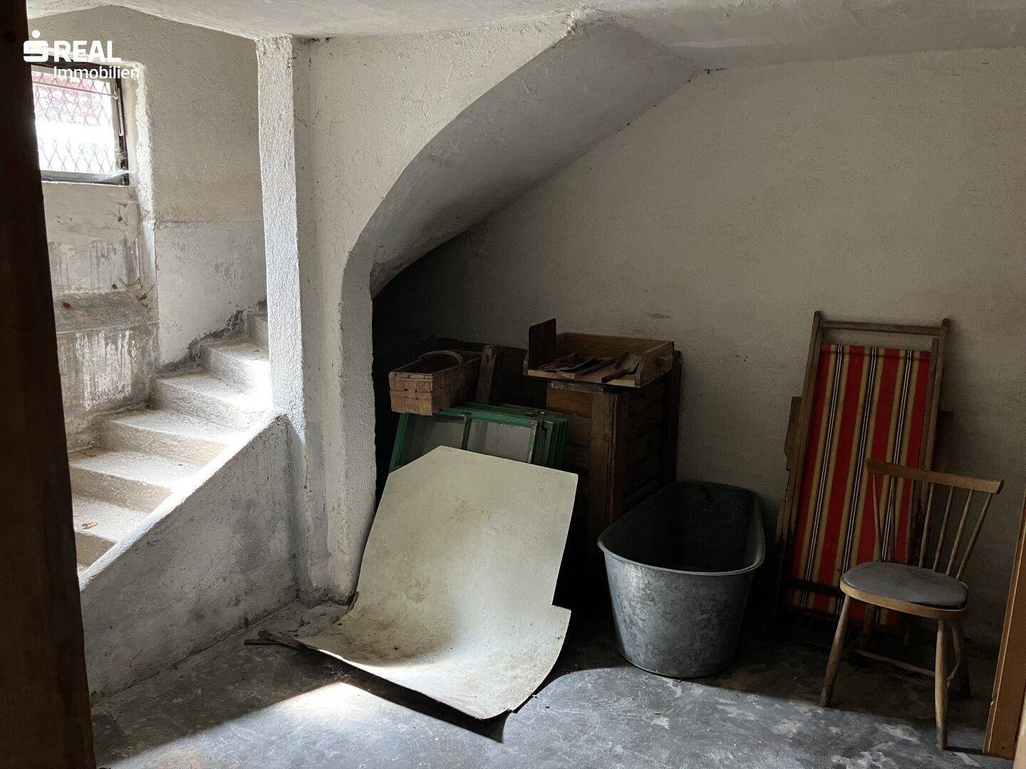 Kellerraum mit Betonwänden und -boden, genutzt als Abstellfläche unter der Treppe.