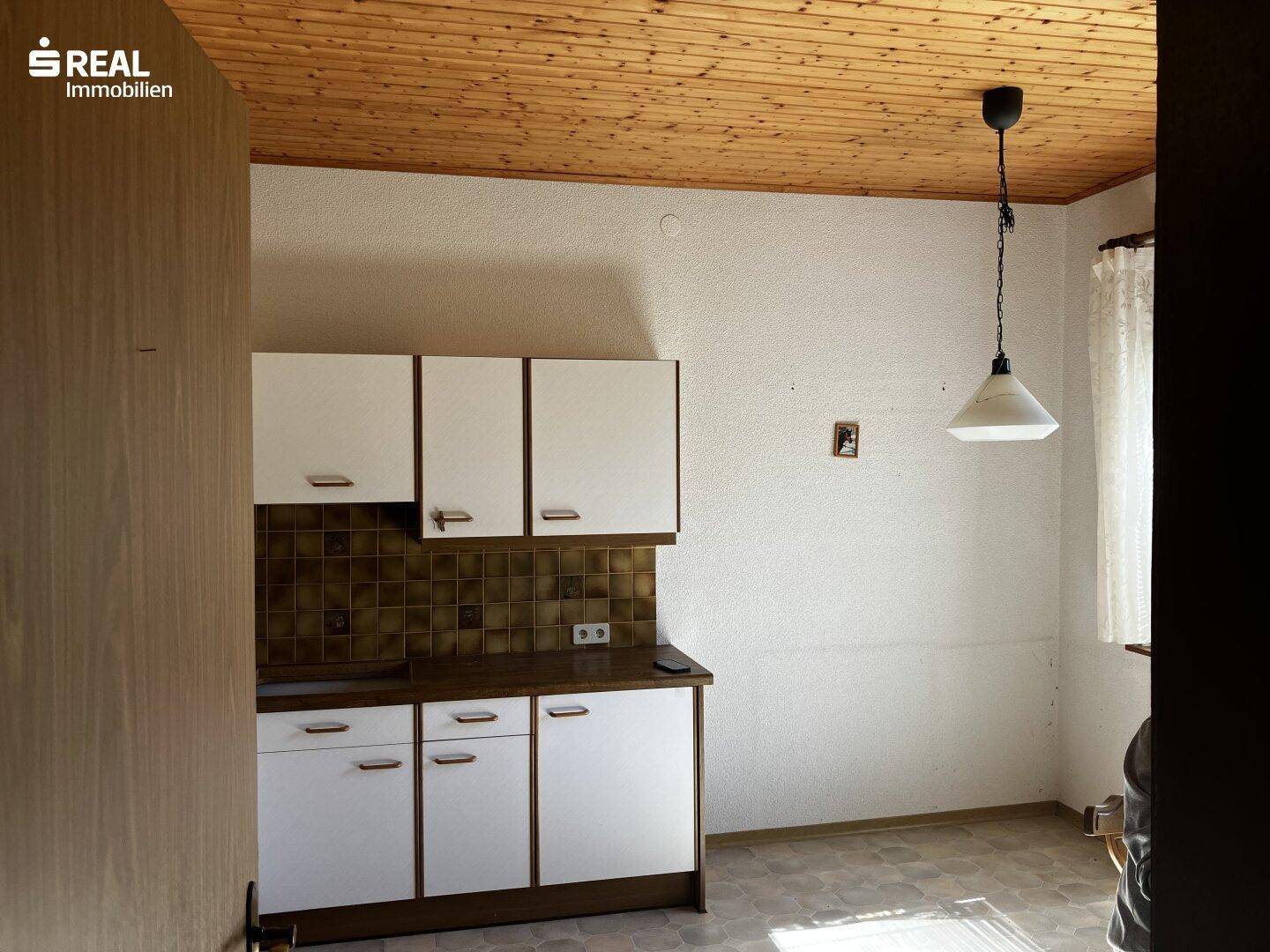Ältere Küche mit weißen Oberschränken, braunen Fliesen und Holzdecke, renovierungsbedürftig.