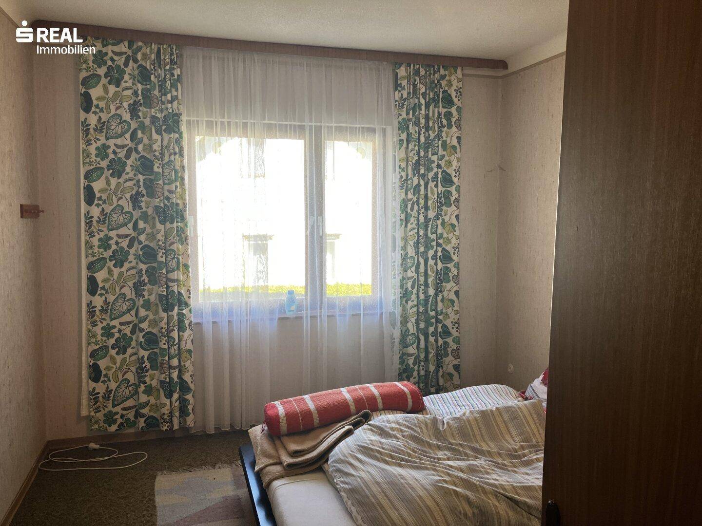 Schlafzimmer mit floralen Vorhängen, Teppichboden und einem Fenster mit Blick ins Grüne.