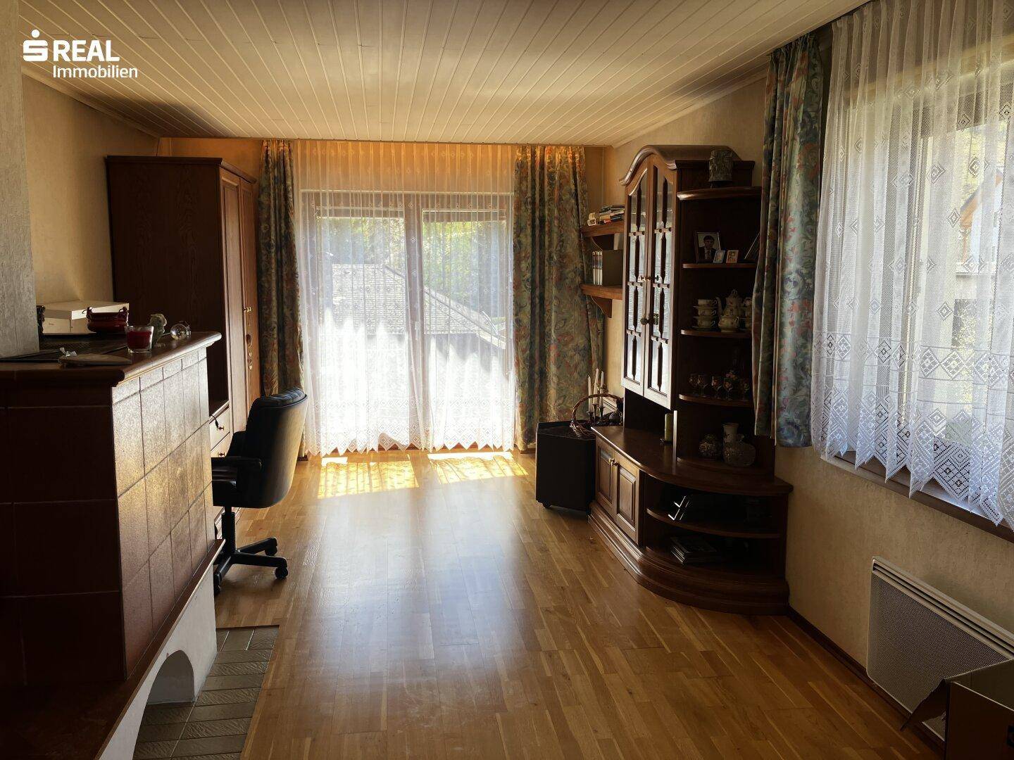 Wohnzimmer mit Holzboden, großem Fenster und Zugang zu einem Außenbereich, inklusive Kachelofen.