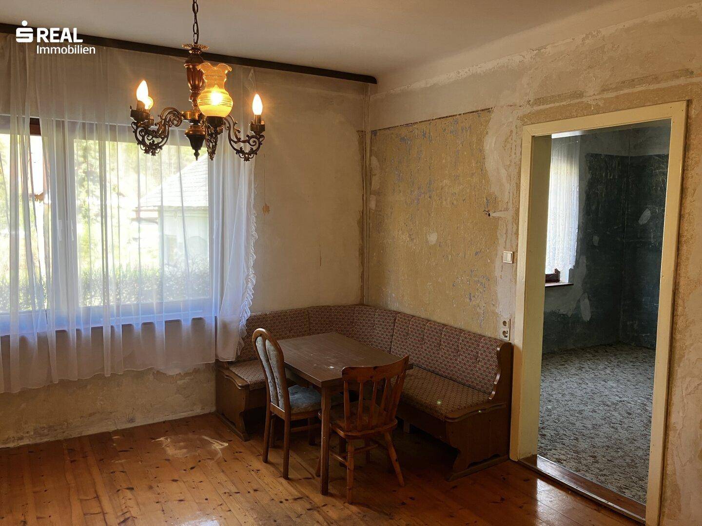 Essbereich mit Eckbank und Holztisch, renovierungsbedürftige Wände und Holzdielenboden.