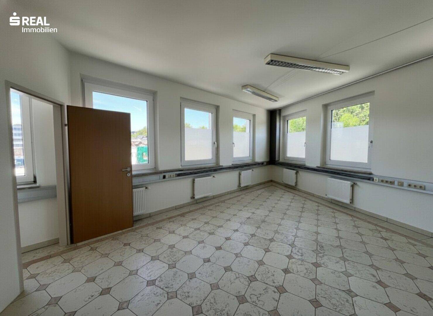 Eckansicht eines hellen Büroraums mit vielen Fenstern und einer braunen Tür.