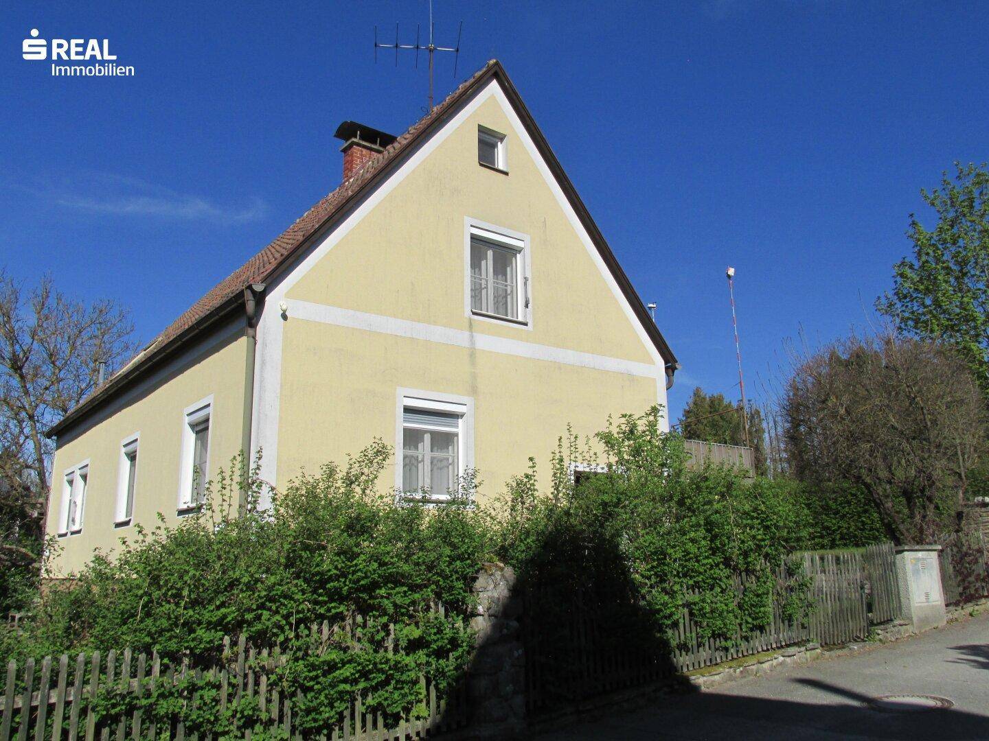 Seitenansicht des Hauses mit gelber Fassade, Satteldach und üppiger Heckenbepflanzung.