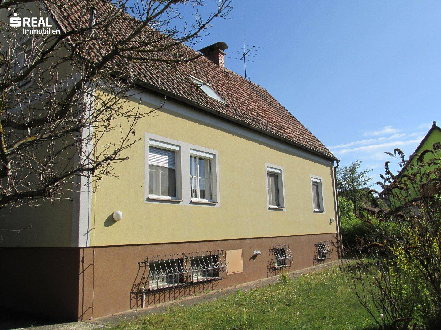 Seitenansicht des Hauses mit gelber Fassade, Satteldach und grüner Umgebung.