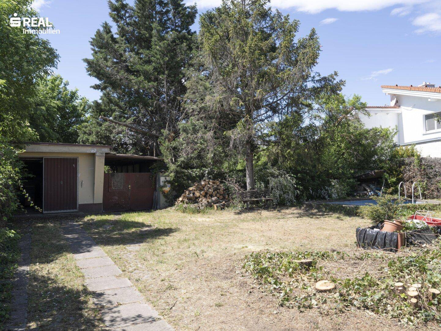 Weitläufiger Garten mit Rasen, hohen Bäumen und einem kleinen Schuppen am Ende des Grundstücks.