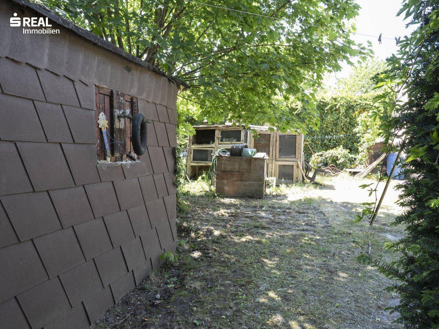 Kleiner Schuppen mit Schindeldach im hinteren Gartenbereich, umgeben von Bäumen und Grün.