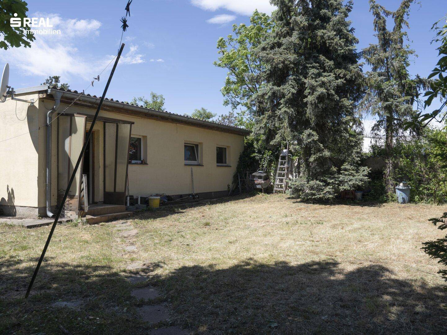 Nebengebäude im Garten mit Fenstern und einer Tür, umgeben von Rasen und hohen Bäumen.