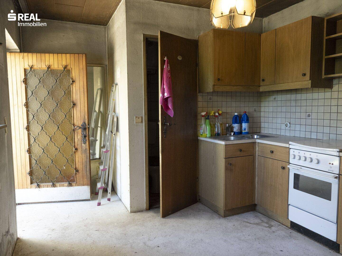 Einfache Küche mit Holzschränken und einem alten Herd, angrenzend an einen weiteren Raum.