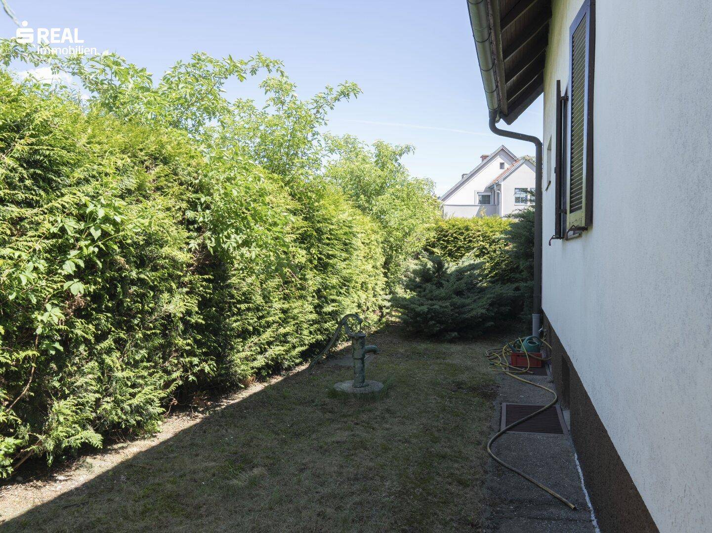 Schmaler Gartenbereich entlang der Hauswand mit hoher Hecke und alter Wasserpumpe.