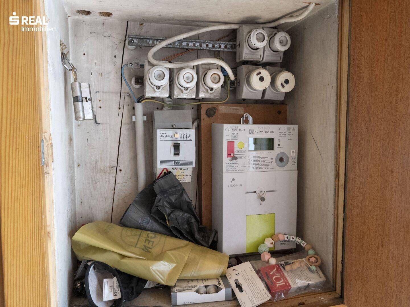 Elektrischer Sicherungskasten und Zähler in einem kleinen Abstellraum mit verschiedenen Gegenständen.