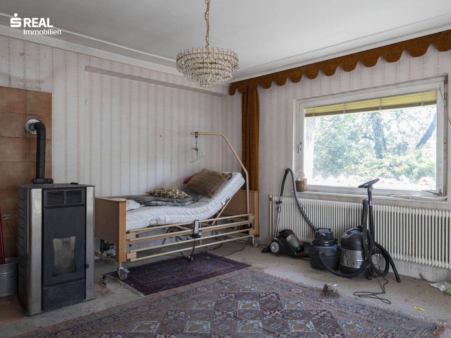 Schlafzimmer mit Pflegebett, Kaminofen und Blick durch das Fenster ins Grüne.