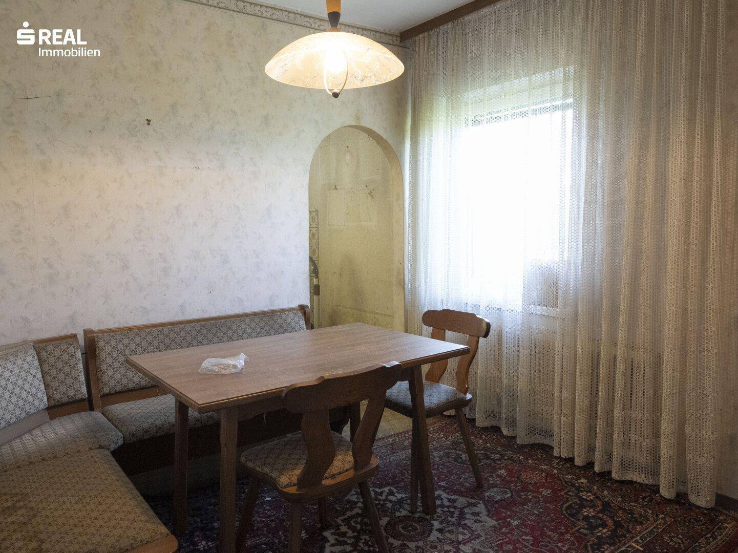 Essbereich mit rustikalem Holztisch, Sitzbank und Fenster mit heller Gardine.
