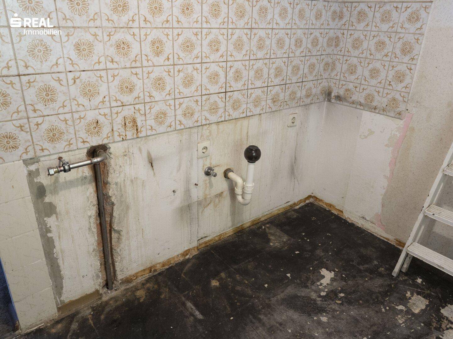Küchenbereich in Sanierung mit sichtbaren Wasseranschlüssen und alten Wandfliesen.