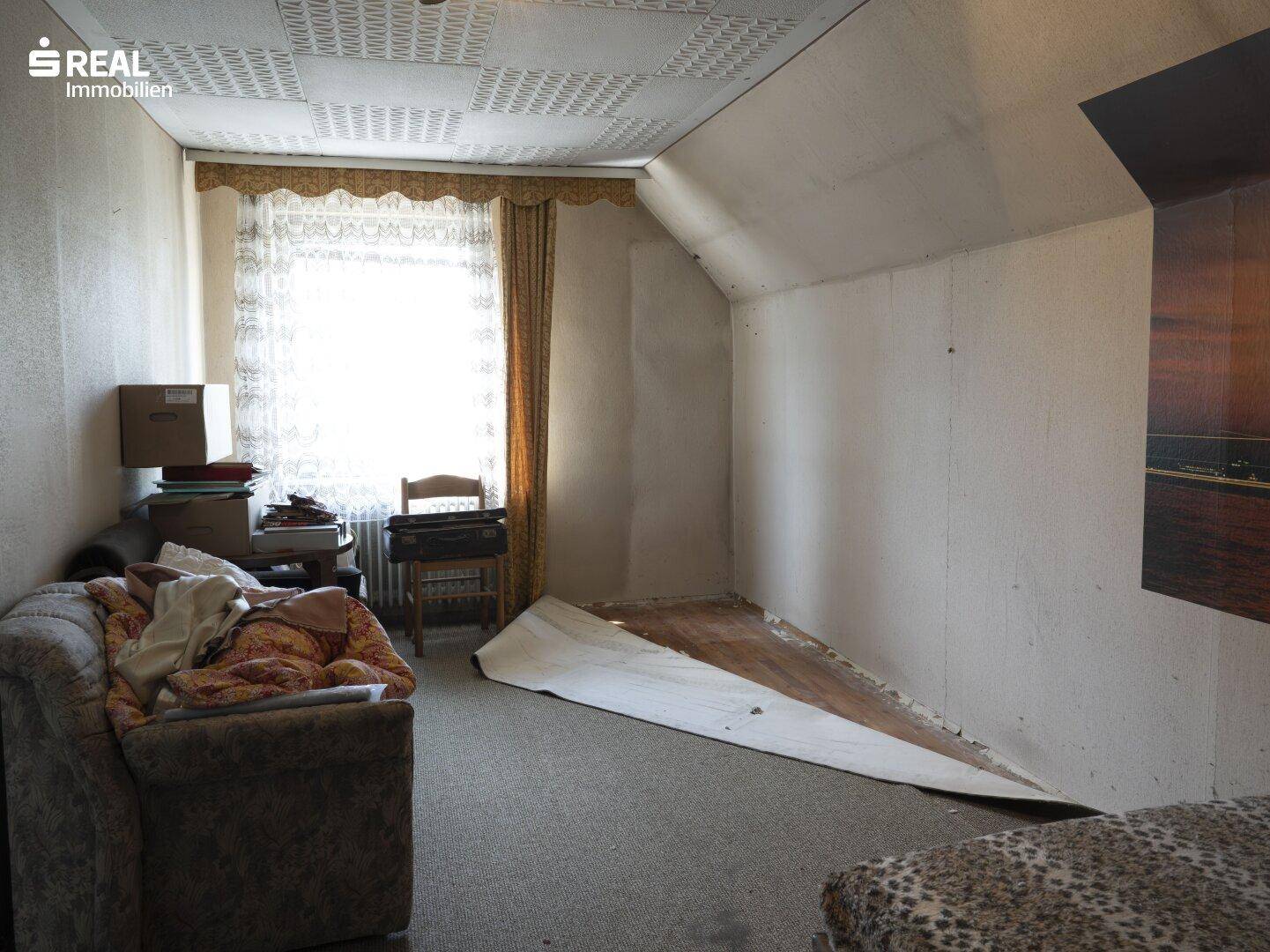 Dachgeschosszimmer mit Sofa, Fenster und schräger Decke, bereit zur Renovierung.