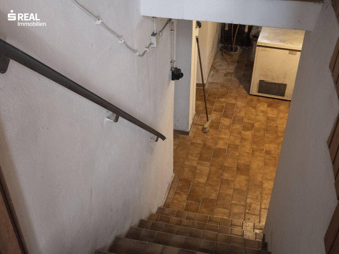 Treppe führt in den Keller mit gefliestem Boden und sichtbaren Versorgungsleitungen.