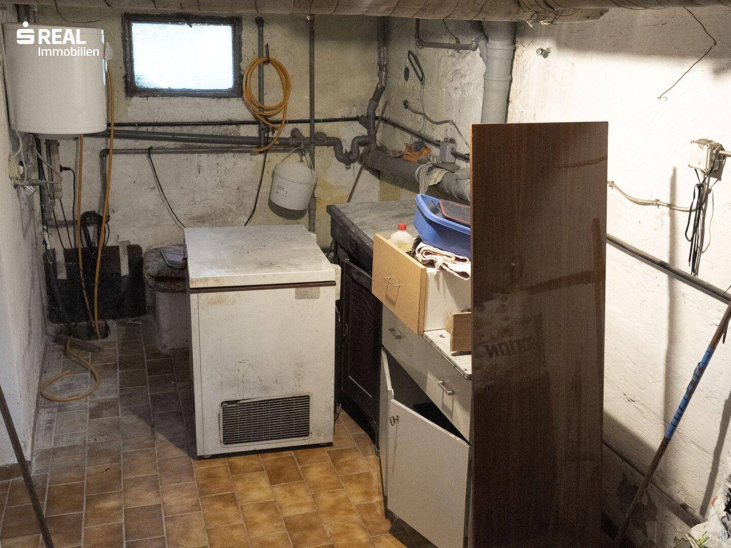 Kellerraum mit Gefriertruhe, Waschbecken und sichtbaren Rohren an der Wand.