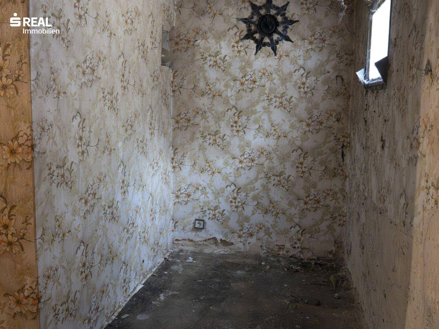 Kleiner Raum mit abblätternder floraler Tapete, abgenutztem Boden und einem kleinen Fenster.