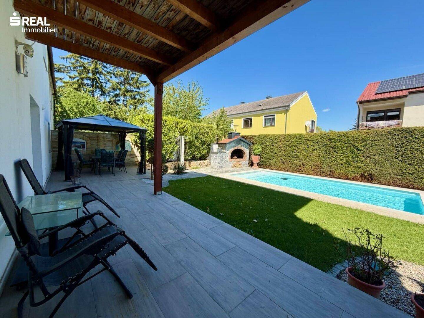 Überdachte Terrasse mit Sitzgelegenheiten, Blick auf den Pool und den gemauerten Grillbereich.