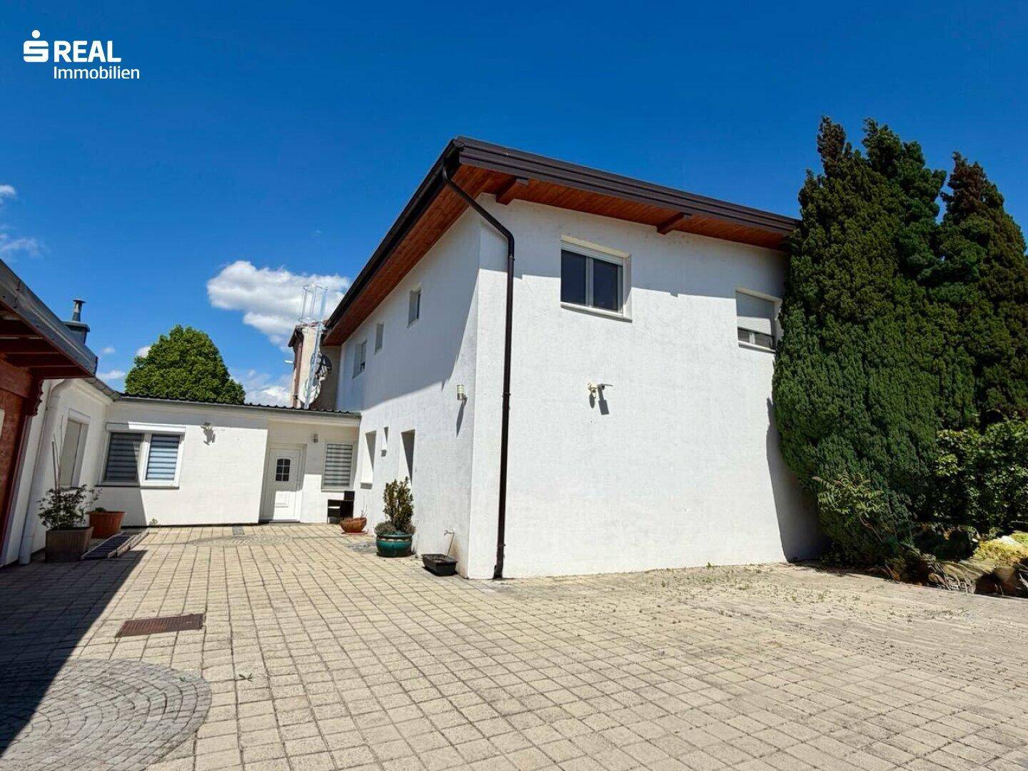 Seitenansicht des weißen Hauses mit gepflastertem Hof und Nebengebäude unter blauem Himmel.