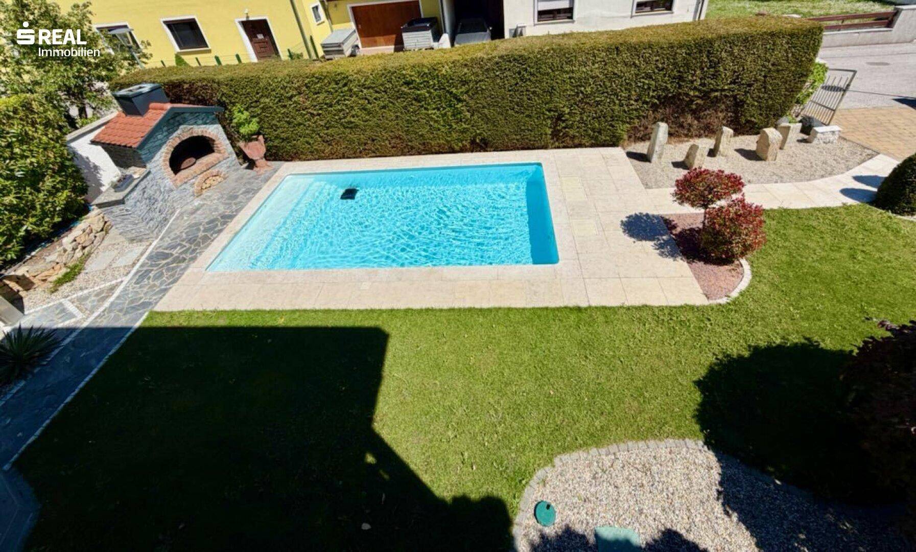 Vogelperspektive des Gartens mit rechteckigem Pool, Rasenfläche und gemauertem Grill.