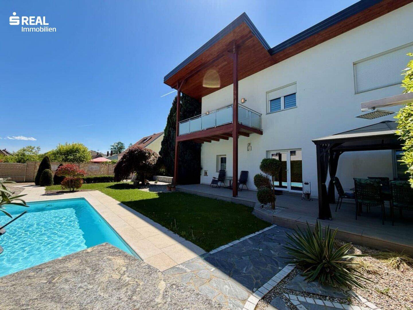 Ansicht des Hauses vom Garten aus mit Pool, Terrasse und Balkon unter blauem Himmel.
