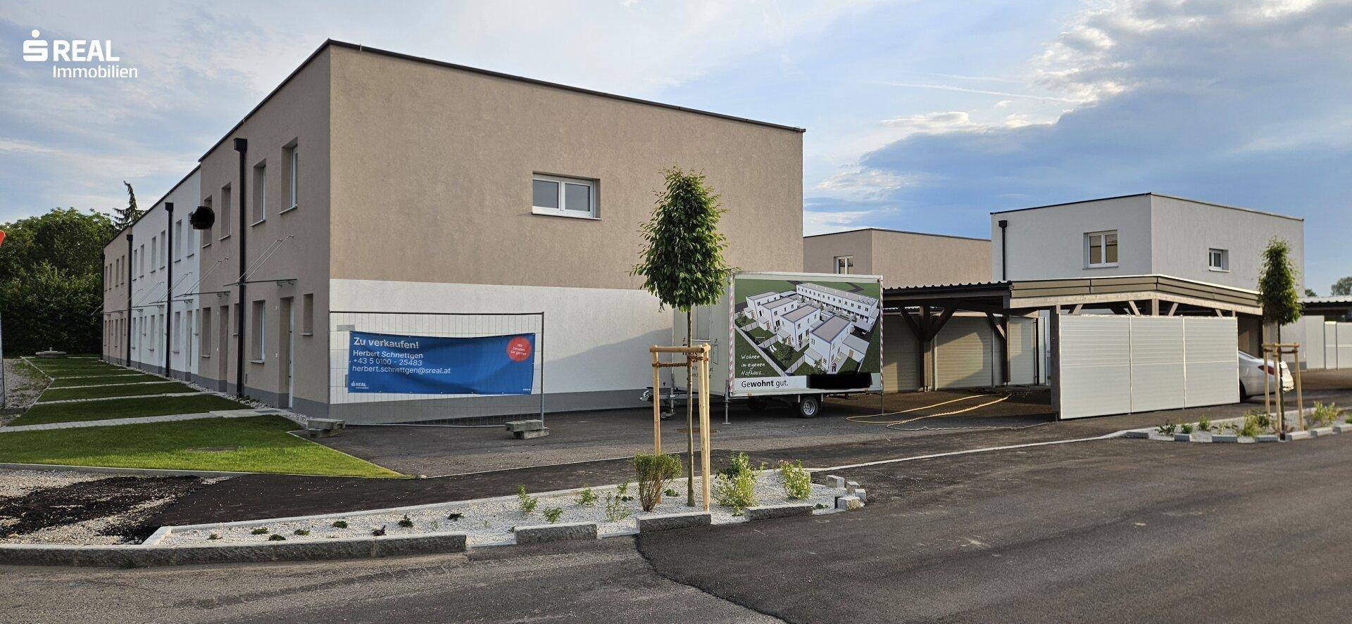 Seitenansicht der Neubauten mit Parkplätzen und einem Informationsbanner zur Immobilie.