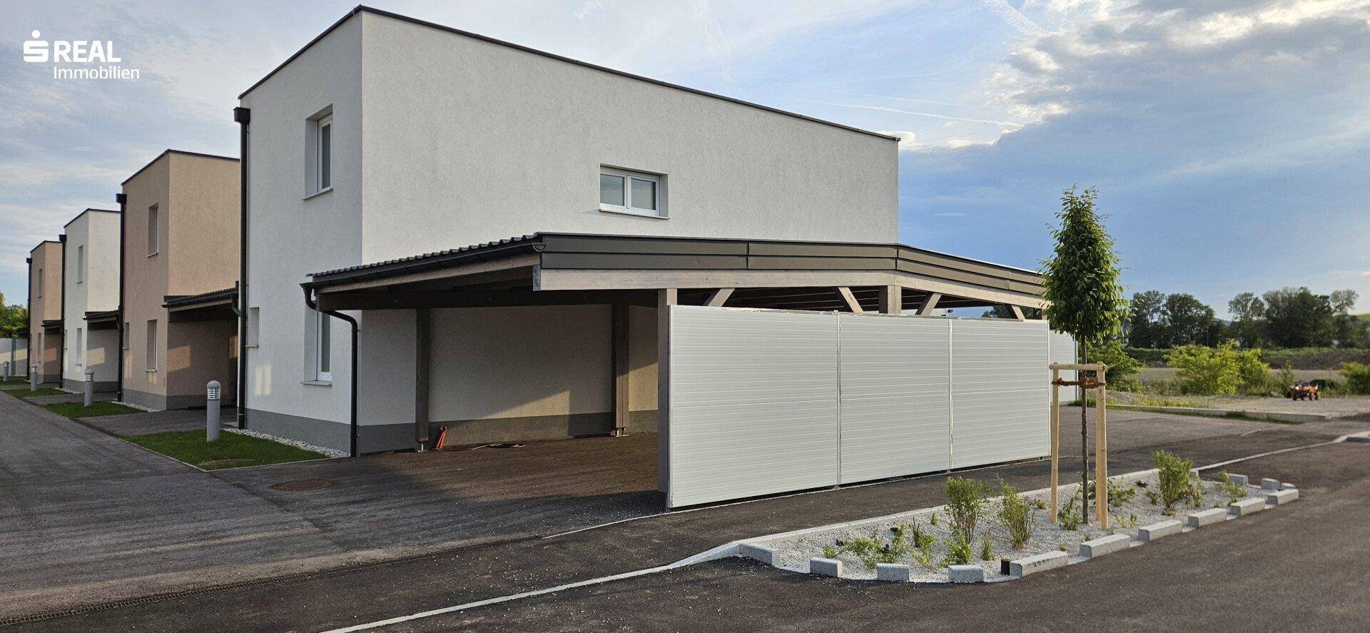 Ansicht eines modernen Reihenhauses mit Carport und Zufahrt, umgeben von ähnlichen Neubauten.