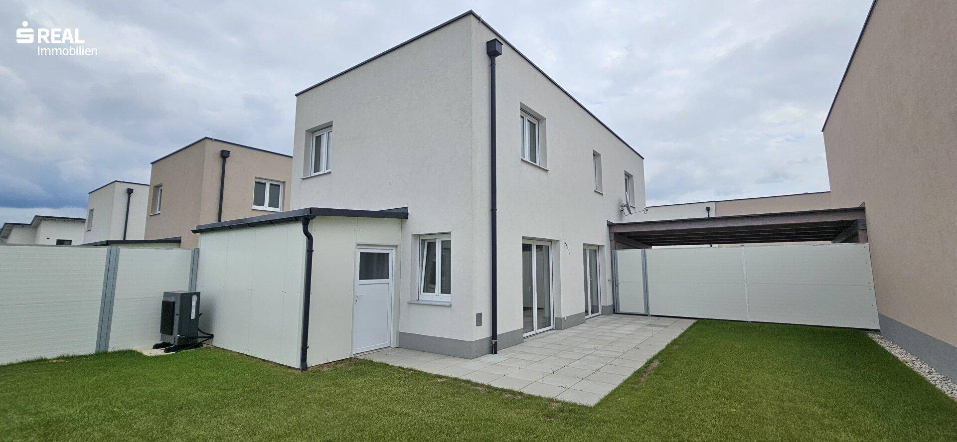Rückseite eines modernen Reihenhauses mit kleiner Terrasse und Rasenfläche unter bewölktem Himmel.