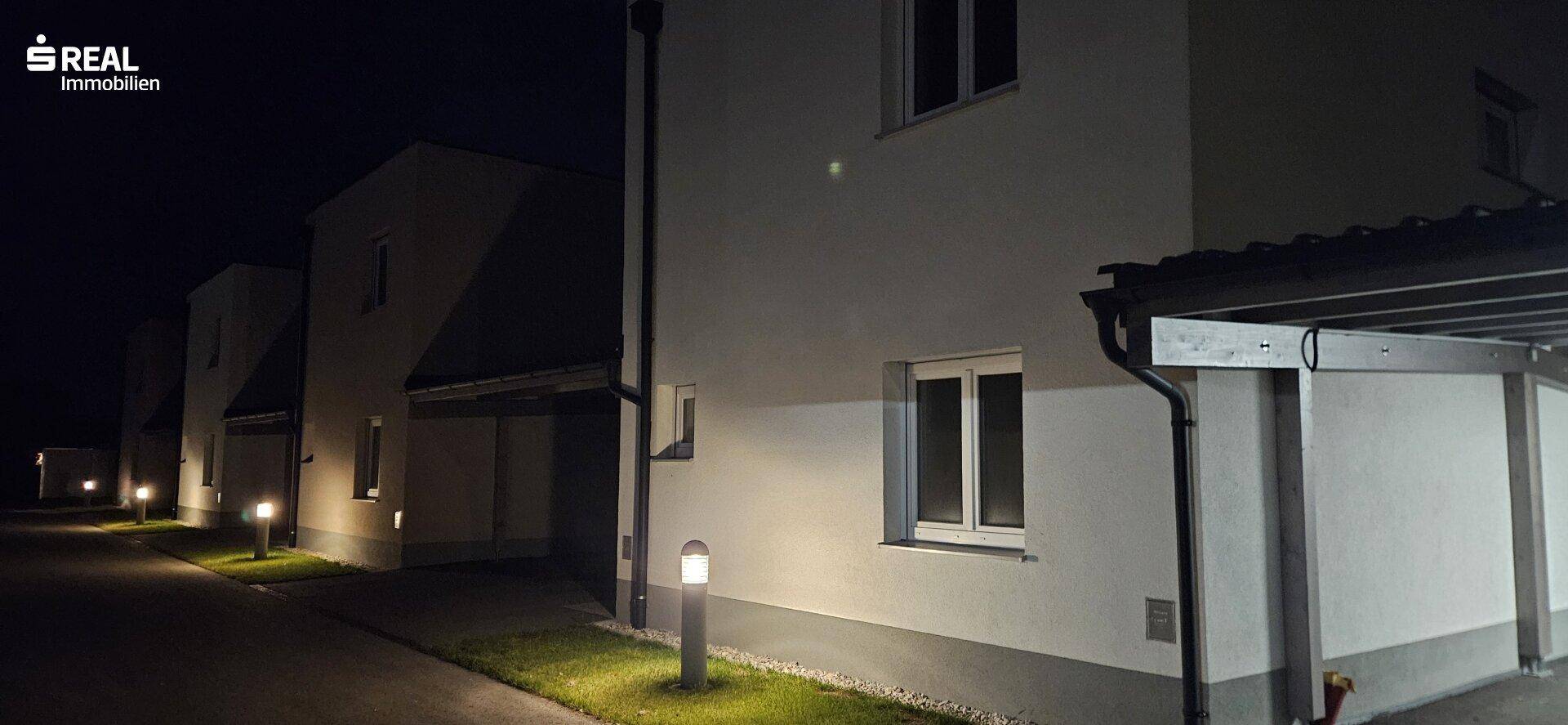 Die modernen Reihenhäuser bei Nacht, beleuchtet von stilvollen Wegleuchten entlang der Zufahrt.