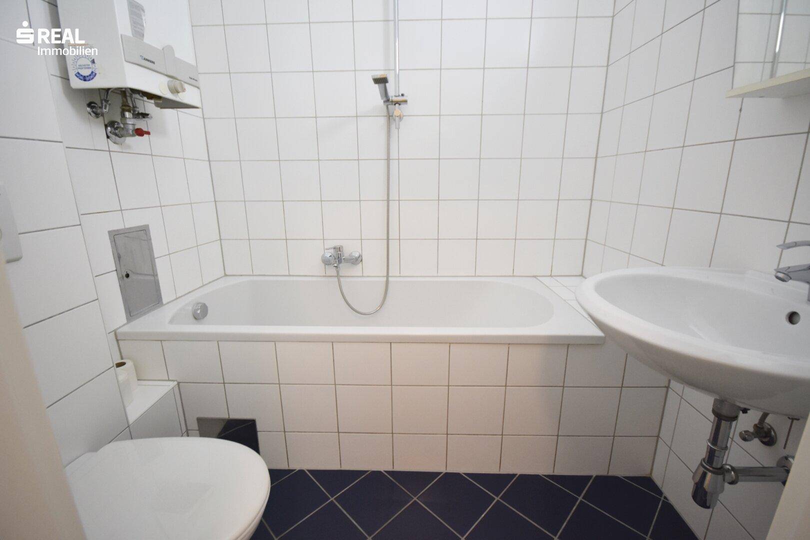 Funktionales Badezimmer mit weiß gefliesten Wänden, dunklem Fliesenboden, Badewanne und WC.