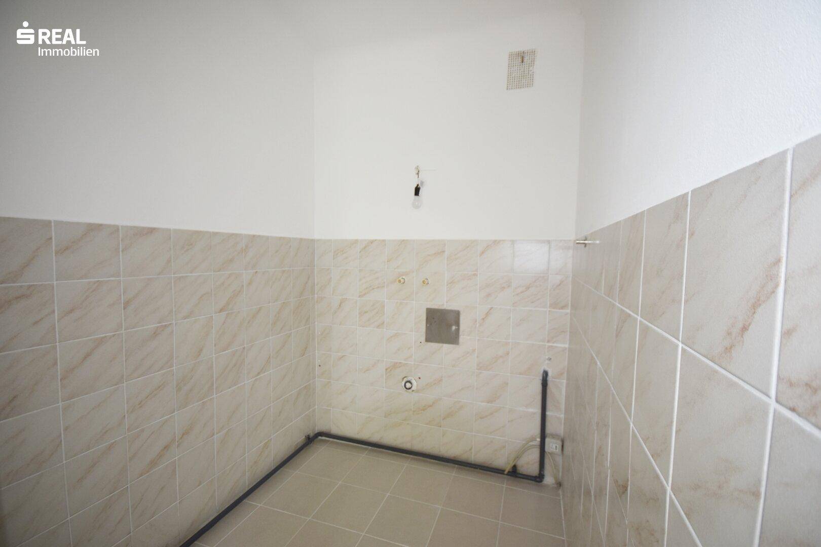 Leerer Raum, vorbereitet für eine Küche, mit halbhohen Fliesenwänden und sichtbaren Anschlüssen.