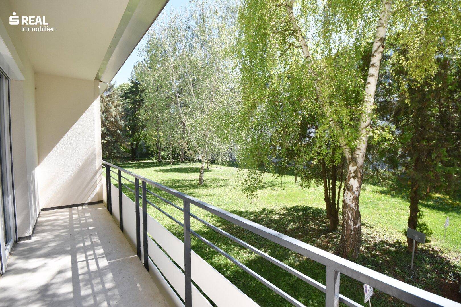 Heller Balkon mit Metallgeländer und weitem Blick auf eine grüne Rasenfläche und Bäume.