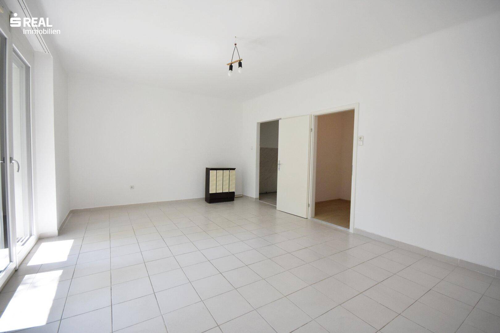 Helles, leeres Wohnzimmer mit Fliesenboden, einem Ofen und Zugang zu einem weiteren Raum.