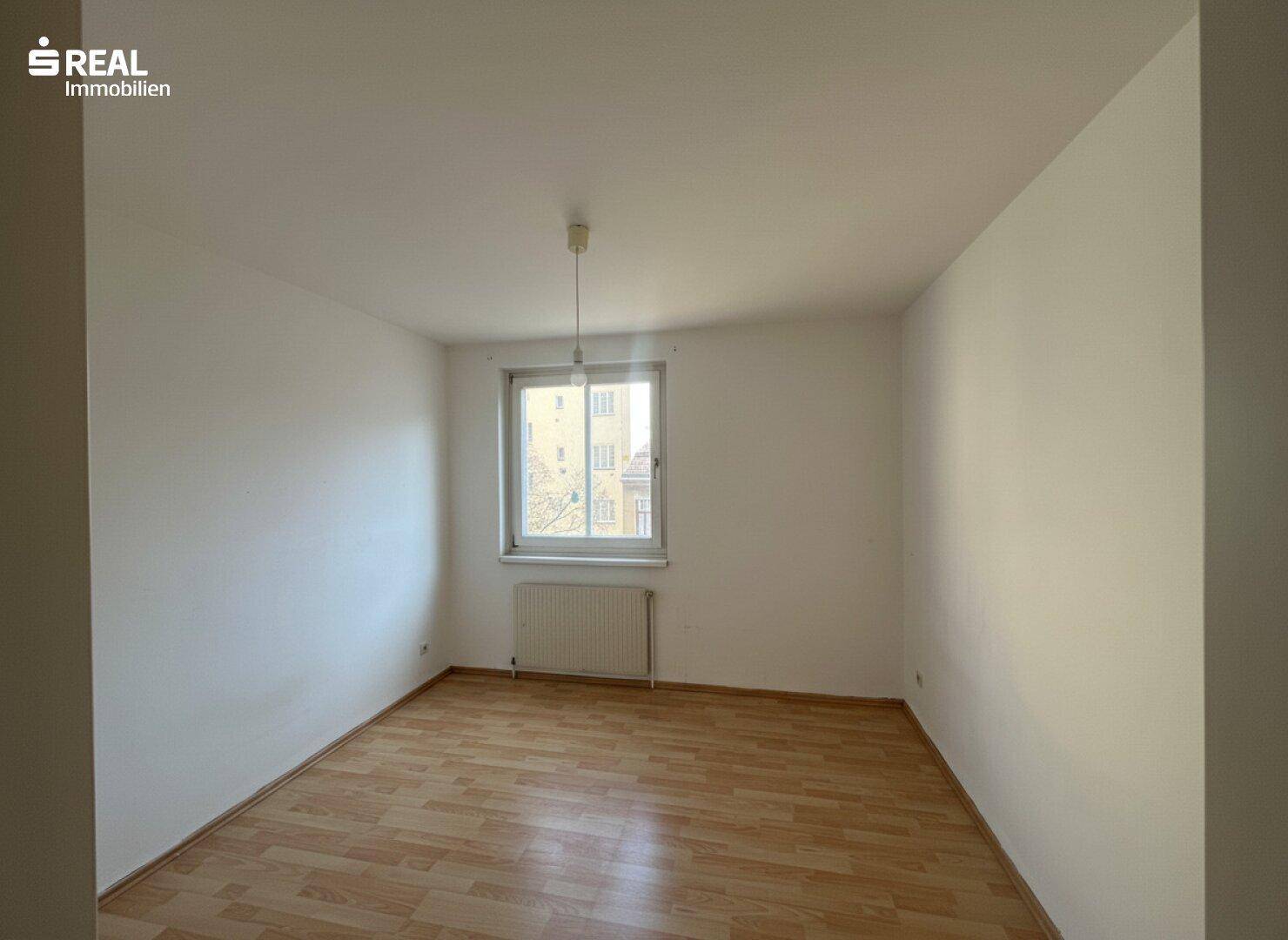 Helles, leeres Schlafzimmer mit Holzboden und einem großen Fenster für viel Tageslicht.