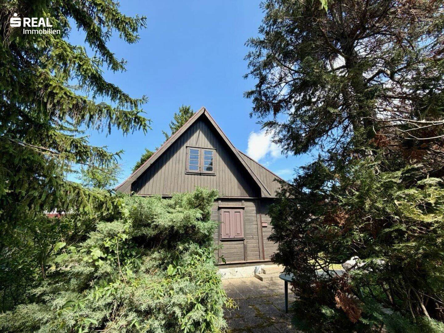 Traditionelles Holzhaus mit Satteldach, umgeben von dichtem Grün und Büschen.