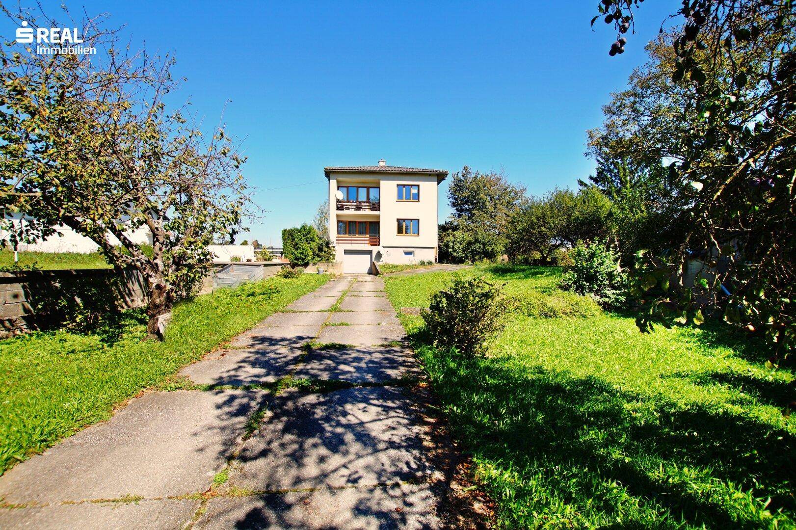 Blick auf das Einfamilienhaus von der Zufahrt, umgeben von viel Grün und Bäumen.