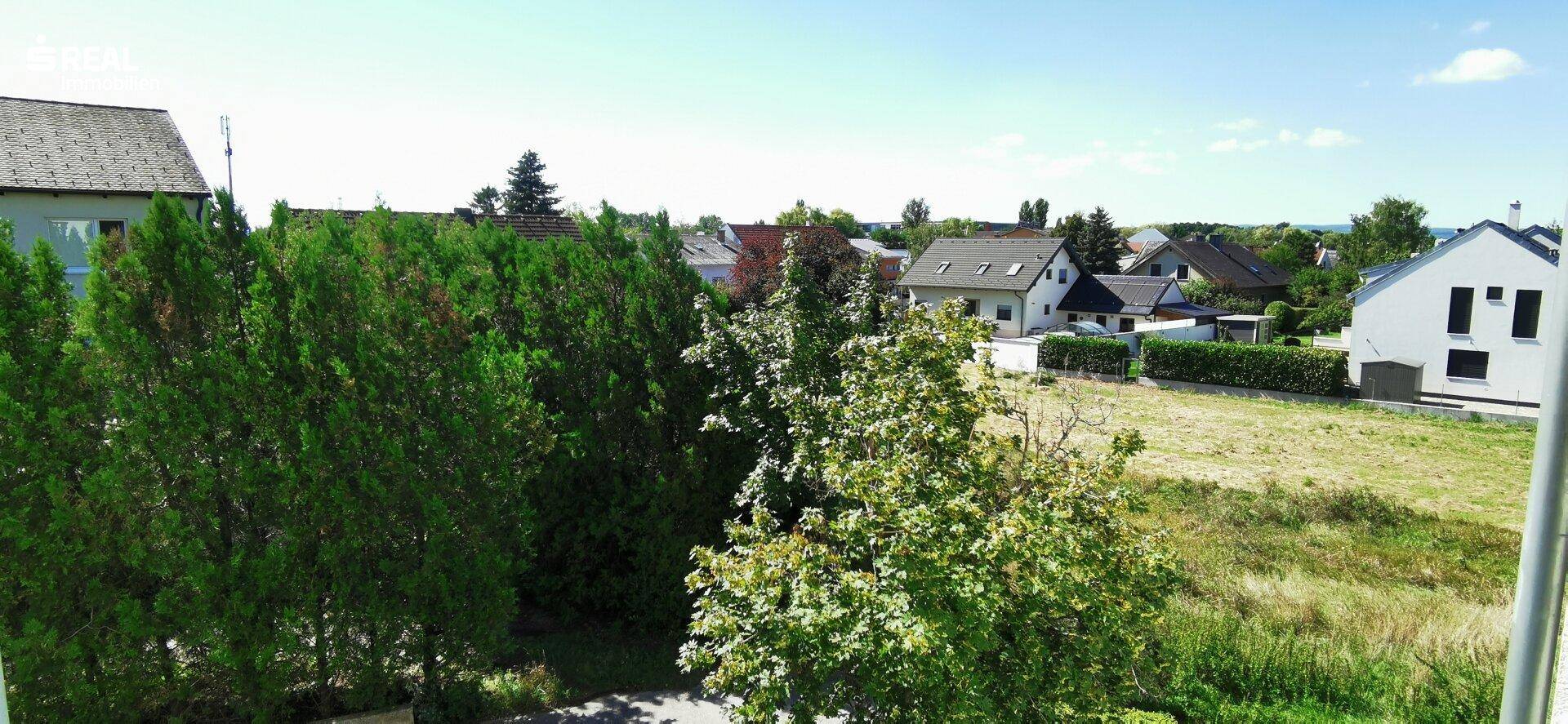 Weitblick über grüne Bäume auf Wohnhäuser und offenes Land unter einem klaren blauen Himmel.