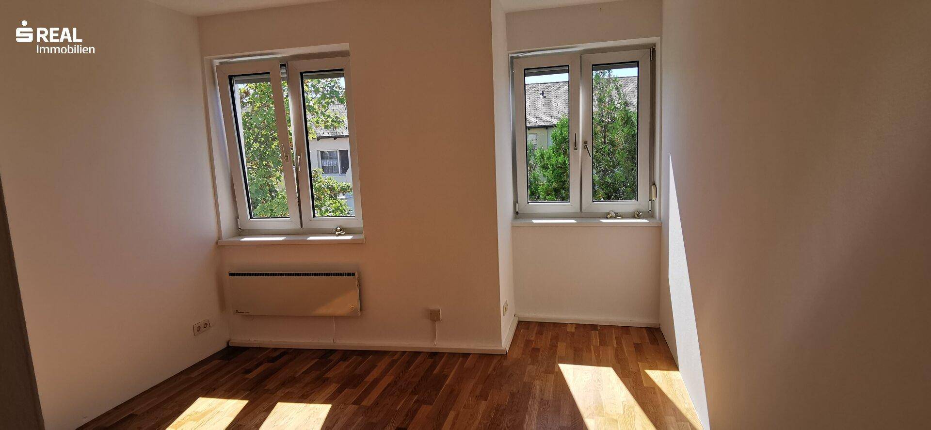 Freundliches Zimmer mit zwei Fenstern, die einen Blick ins Grüne bieten, und hellem Holzfußboden.