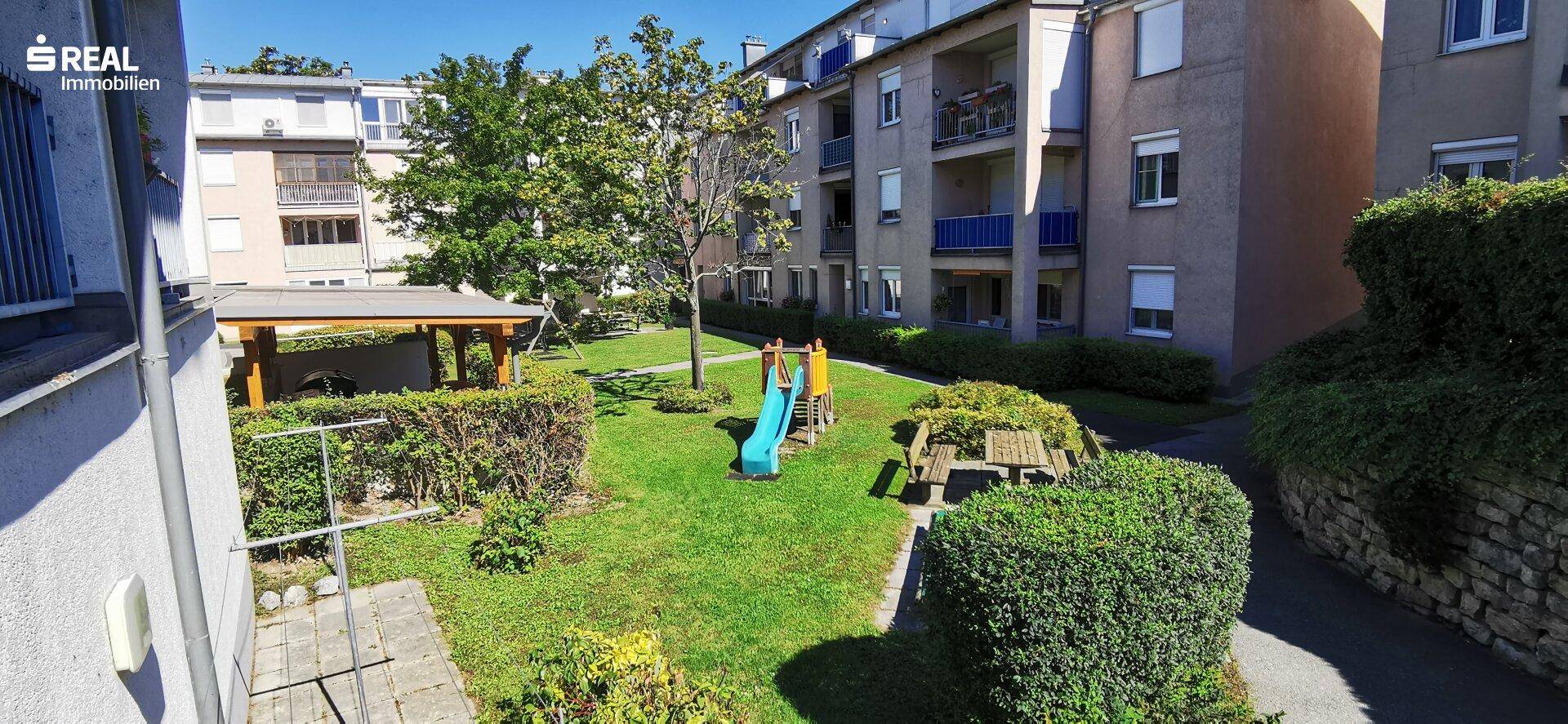 Grüner Innenhof einer Wohnanlage mit Rasenfläche, Bäumen und einem kleinen Spielplatz für Kinder.