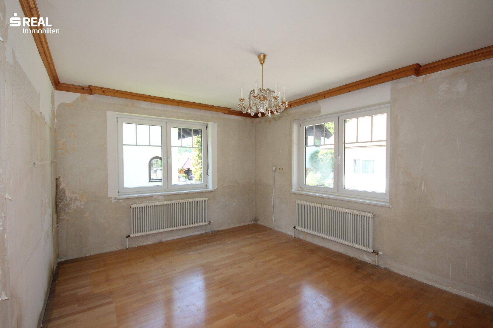 Helles Wohnzimmer mit Parkettboden, zwei Fenstern und einem eleganten Kronleuchter.